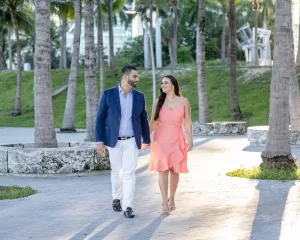 Professional Photographer A couple walks hand-in-hand on a palm tree-lined path, smiling at each other. Perfect for wedding photography, the man wears a blue blazer and white pants, while the woman’s peach dress glows in the sunny, green setting. www.maranonimagery.com