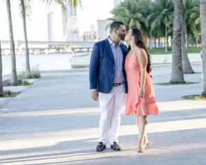 Professional Photographer A couple stands outdoors by the water, surrounded by palm trees, sharing a kiss. The man wears a blue blazer and white pants, while the woman in a coral dress creates a romantic scene perfect for wedding photography. City buildings are visible in the background. www.maranonimagery.com