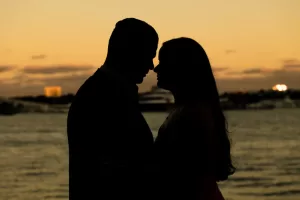 Professional Photographer Silhouette of a couple facing each other closely by the water at sunset, with a soft orange and yellow sky in the background—an intimate portrait capturing romance in golden hour light. www.maranonimagery.com