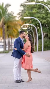 Professional Photographer A couple kisses on a tree-lined pathway at sunset, captured in stunning wedding photography. The woman lifts one leg in a coral dress and heels, while the man wears a blue blazer and light pants under softly curving streetlights. www.maranonimagery.com