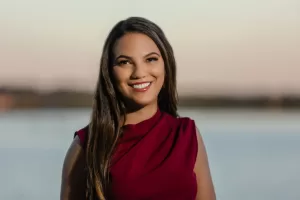 Professional Photographer A woman with long brown hair smiles while wearing a sleeveless burgundy top, standing outdoors near calm water at sunset—an ideal setting for stunning portraits or elegant headshots. www.maranonimagery.com
