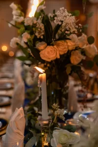 Professional Photographer A lit white candle in a glass holder sits on a decorated dinner table with white flowers, green leaves, and napkins folded upright, creating an elegant and romantic atmosphere perfect for wedding photography. www.maranonimagery.com