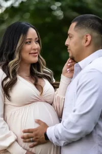 Professional Photographer A pregnant woman in a light dress smiles and gently touches the face of a man who is touching her belly. They stand close together outdoors, greenery behind them—a moment as intimate and timeless as classic wedding photography. www.maranonimagery.com