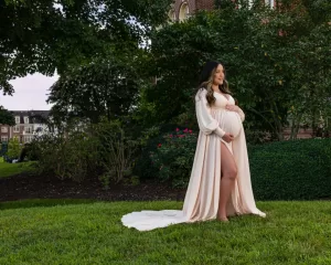 Professional Photographer A pregnant woman in a long, flowing cream dress stands barefoot on grass in a garden, gently holding her belly and smiling. Lush greenery and a brick building create the perfect backdrop for timeless maternity portraits. www.maranonimagery.com