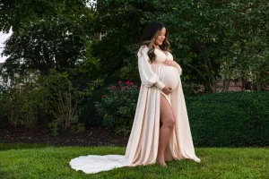 Professional Photographer A pregnant woman in a flowing, light-colored gown stands on grass in a garden, cradling her belly and smiling thoughtfully. Lush greenery and blooming bushes surround her, creating a serene setting perfect for portraits. www.maranonimagery.com