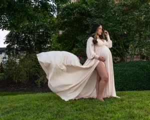 Professional Photographer A pregnant woman stands barefoot on grass in a garden, wearing a flowing, long cream-colored dress that billows behind her. She gently touches her hair and cradles her belly, as a photographer captures the moment amid lush greenery. www.maranonimagery.com