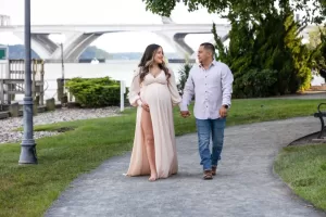 Professional Photographer A pregnant woman in a flowing, light pink dress walks hand-in-hand with a man in a white shirt and jeans along a riverside path, capturing the feel of wedding photography with portraits against a scenic bridge and greenery backdrop. www.maranonimagery.com