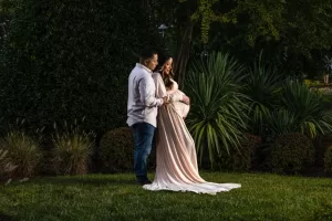 Professional Photographer A couple stands together outdoors on grass, the man gently holding the woman from behind as she cradles her pregnant belly—a beautiful moment captured by a skilled photographer, both dressed in light clothing and surrounded by lush greenery. www.maranonimagery.com