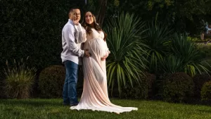 Professional Photographer A couple poses outdoors; the pregnant woman wears a long, flowing pale pink dress while the man stands beside her in a light shirt and jeans. Captured by a photographer, they are surrounded by lush green plants and grass. www.maranonimagery.com