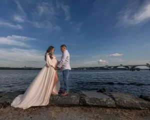Professional Photographer A couple stands on rocks by the water, holding hands and facing each other in a moment captured by a skilled photographer. The woman, appearing pregnant in her flowing dress, stands beneath a blue sky with wispy clouds and a bridge in the background. www.maranonimagery.com