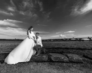 Professional Photographer A man kneels in front of a pregnant woman in a long dress by a riverside, holding her belly. The dramatic sky and distant bridge set the mood, giving this black-and-white photo a timeless look reminiscent of classic wedding photography. www.maranonimagery.com