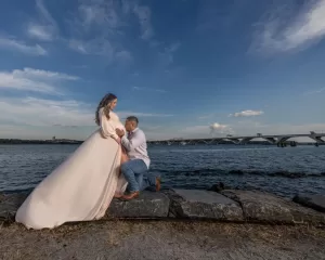 Professional Photographer A man kneels by a pregnant woman in a flowing pale dress, gently touching her belly. Outdoors by the water, with a bridge and blue sky in the background, this moment captures true love—perfect for maternity portraits. www.maranonimagery.com