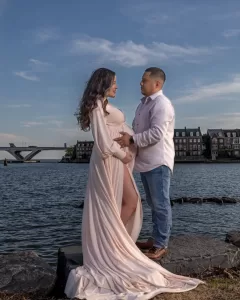 Professional Photographer A photographer captures a pregnant woman in a flowing light pink dress standing on rocks by the water, facing a man in a white shirt and jeans. They gaze at each other, with townhouses and a bridge in the background under a blue sky. www.maranonimagery.com
