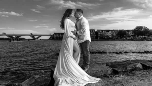 Professional Photographer A couple stands on rocks by the water, embracing and gazing at each other in a romantic wedding photography portrait. The woman’s long, flowing dress with a slit and the scenic background create an unforgettable moment under the cloudy sky. www.maranonimagery.com