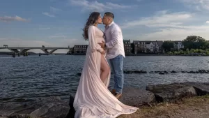 Professional Photographer A couple stands on rocks by the water, facing each other and touching the woman’s pregnant belly. Perfect for portraits, the woman wears a flowing light pink dress; the man wears jeans and a white shirt. Houses and a bridge are visible in the background. www.maranonimagery.com