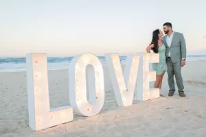Professional Photographer A couple stands on a sandy beach beside large, illuminated LOVE letters. Captured by a skilled photographer, they smile and lean affectionately toward each other, framed by the ocean and sky in the background. www.maranonimagery.com