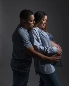 Professional Photographer A man gently hugs a pregnant woman from behind as they share a peaceful moment. The woman holds her exposed belly, creating a portrait of love and anticipation against a dark, plain background. www.maranonimagery.com