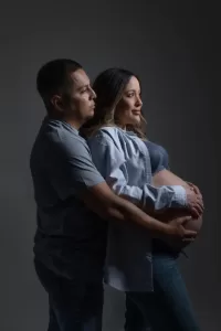 Professional Photographer A man stands behind a pregnant woman, both gently holding her belly and looking forward with soft expressions. Dressed casually, they pose for heartfelt portraits against a dark, neutral background. www.maranonimagery.com