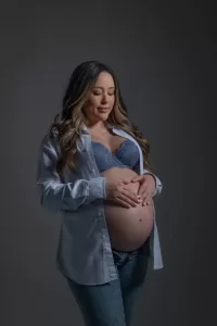 Professional Photographer A pregnant woman in a blue bra and unbuttoned shirt stands against a plain background, gently cradling her bare belly and smiling softly with her eyes closed—a beautiful portrait capturing a tender moment. www.maranonimagery.com