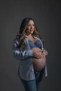 Professional Photographer A pregnant woman with long hair stands against a gray background, wearing an open button-down shirt, a blue bralette, and jeans. The photographer captures her gently cradling her bare baby bump and smiling softly in this beautiful portrait. www.maranonimagery.com