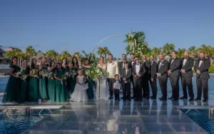 Professional Photographer A large wedding party stands outdoors on a clear platform over water, with bridesmaids in green dresses, groomsmen in black suits, and the bride and groom in the center holding bouquets—captured perfectly by the photographer. Palm trees and blue sky complete the scene. www.maranonimagery.com
