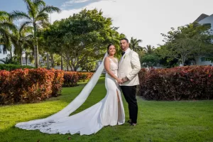 Professional Photographer A bride and groom stand together on a green lawn, surrounded by lush trees and bushes. Perfect for wedding photography, the bride wears a long white dress with a train, and the groom wears a cream blazer and black pants as they look at the camera. www.maranonimagery.com