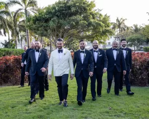 Professional Photographer A groom in a white tuxedo jacket walks on grass with six smiling groomsmen in black tuxedos, surrounded by trees and tropical plants, creating a portrait-worthy moment. www.maranonimagery.com
