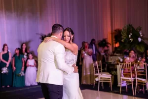 Professional Photographer A bride and groom share their first dance at a wedding reception, surrounded by guests in elegant attire. Captured by a skilled wedding photographer, this portrait highlights the bride’s joy against decorated tables and lush greenery. www.maranonimagery.com
