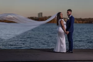 Professional Photographer A bride and groom stand hand in hand by the water at sunset, captured in stunning wedding photography. The bride’s veil flows in the wind as they look toward the camera, framed by a city skyline and elegant wedding attire. www.maranonimagery.com