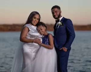 Professional Photographer A woman in a white dress, a young girl in a white and blue dress, and a man in a blue suit stand together by the water, smiling at the camera during sunset—captured perfectly by a skilled wedding photography professional. www.maranonimagery.com