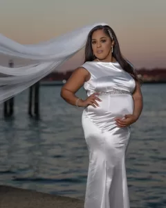Professional Photographer A pregnant woman in a white satin gown poses by the water at sunset, holding her belly and wearing a long, flowing veil—capturing the elegance found in wedding photography. www.maranonimagery.com