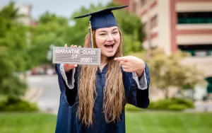 Professional Photographer A smiling graduate in a navy cap and gown holds a sign reading Quarantine Class of 2020 and points at it while standing outdoors, as a photographer captures the moment with trees and buildings in the background. www.maranonimagery.com