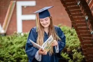 Professional Photographer A young woman wearing a navy blue graduation cap and gown smiles while reading a book outdoors, standing near a brick arch with greenery and a large white letter F in the background—perfect for graduation headshots. www.maranonimagery.com