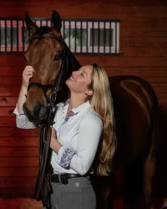 Professional Photographer A woman with long blonde hair stands in a horse stable, smiling as she gently holds a brown horse’s head close—capturing the warmth perfect for pet photography. The background features wood paneling and barred windows. www.maranonimagery.com