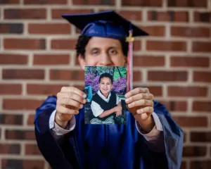 Professional Photographer A photographer captures a person in a blue graduation gown and cap holding up a childhood photo, smiling in front of a brick wall. The focus is on the photo, with the graduate’s face artistically blurred in the background. www.maranonimagery.com