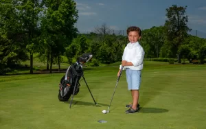 Professional Photographer A young boy in a white shirt and light blue shorts stands on a golf course, smiling and holding a golf club near a hole, striking a pose as if ready for playful headshots, with his golf bag beside him and trees in the background. www.maranonimagery.com