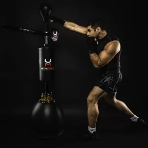 Professional Photographer A man wearing black athletic gear punches a tall, freestanding boxing trainer with extended padded arms and a speed bag, against a dark background—captured by a skilled photographer. www.maranonimagery.com