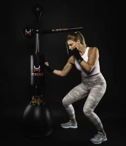 Professional Photographer A woman in athletic wear punches a black standing boxing trainer with moving arms in a dark studio setting. Captured by the photographer, she is in an active stance, wearing gloves, a white crop top, and light camo leggings. www.maranonimagery.com
