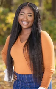 Professional Photographer A young woman with long straight hair, wearing an orange sweater and blue plaid skirt, smiles outdoors with one hand on her hip. Perfect for portraits or headshots, the background features blurred greenery and sunlight. www.maranonimagery.com