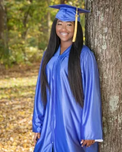 Professional Photographer A woman wearing a blue graduation cap and gown stands and smiles while leaning against a tree in an outdoor setting, as a photographer captures the moment amid fallen leaves and green foliage in the background. www.maranonimagery.com