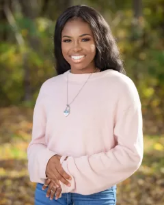 Professional Photographer A woman wearing a light pink sweater and a pendant necklace stands outdoors, smiling, with a blurred background of trees and fall foliage—perfect for natural headshots or portraits. www.maranonimagery.com