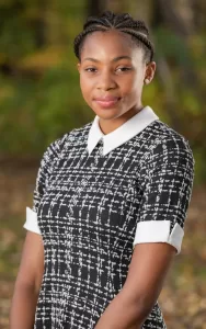 Professional Photographer A young woman with braided hair, wearing a black and white patterned dress with a white collar and cuffs, stands outdoors with a soft smile—captured in stunning detail by a talented headshots photographer against a blurred green and brown background. www.maranonimagery.com