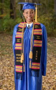 Professional Photographer A smiling graduate wearing a blue cap and gown, a kente stole with Potomac Valley Section and NCNW in gold letters, stands outdoors on a path covered with fallen leaves—perfect for headshots capturing this milestone moment. www.maranonimagery.com