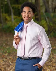 Professional Photographer A young man stands outdoors, smiling confidently. Dressed in a light pink dress shirt, pink tie, and navy pants with a blue jacket over his shoulder, he radiates style—an ideal moment for creative portraits amid the wooded area’s green and yellow leaves. www.maranonimagery.com
