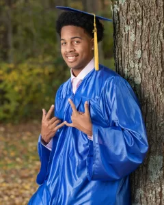Professional Photographer A young man in a blue graduation gown and cap leans against a tree, smiling and making hand signs. Captured by a photographer, this portrait highlights his joy amidst green foliage and fallen leaves outdoors. www.maranonimagery.com