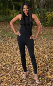 Professional Photographer A woman stands outdoors on fallen autumn leaves, smiling with hands on hips. She wears a sleeveless black jumpsuit and silver heels. Perfect for photographer headshots, the scene features her long hair and trees with green and yellow leaves in the background. www.maranonimagery.com