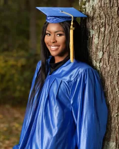 Professional Photographer A young woman wearing a shiny blue graduation gown and cap with a yellow tassel smiles while leaning against a tree outdoors, perfect for headshots by a professional photographer. www.maranonimagery.com