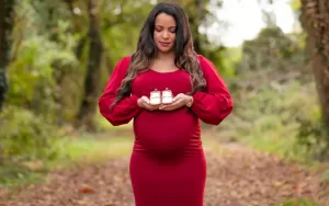 Professional Photographer A pregnant woman in a long red dress stands on a forest path, holding baby shoes and looking down at them. The green, blurred background with trees and fallen leaves creates a serene setting for this beautiful maternity portrait. www.maranonimagery.com