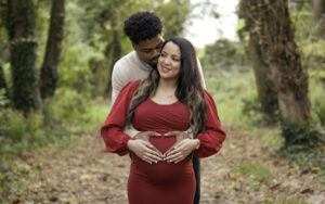 Professional Photographer A man stands behind a pregnant woman in a red dress, both holding her belly and smiling, while standing on a leafy path in a wooded area—an intimate moment perfect for portraits. www.maranonimagery.com