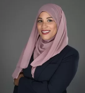 Professional Photographer A woman wearing a mauve hijab and a dark blazer, who is a talented photographer, smiles confidently with her arms crossed in front of a neutral gray background. www.maranonimagery.com